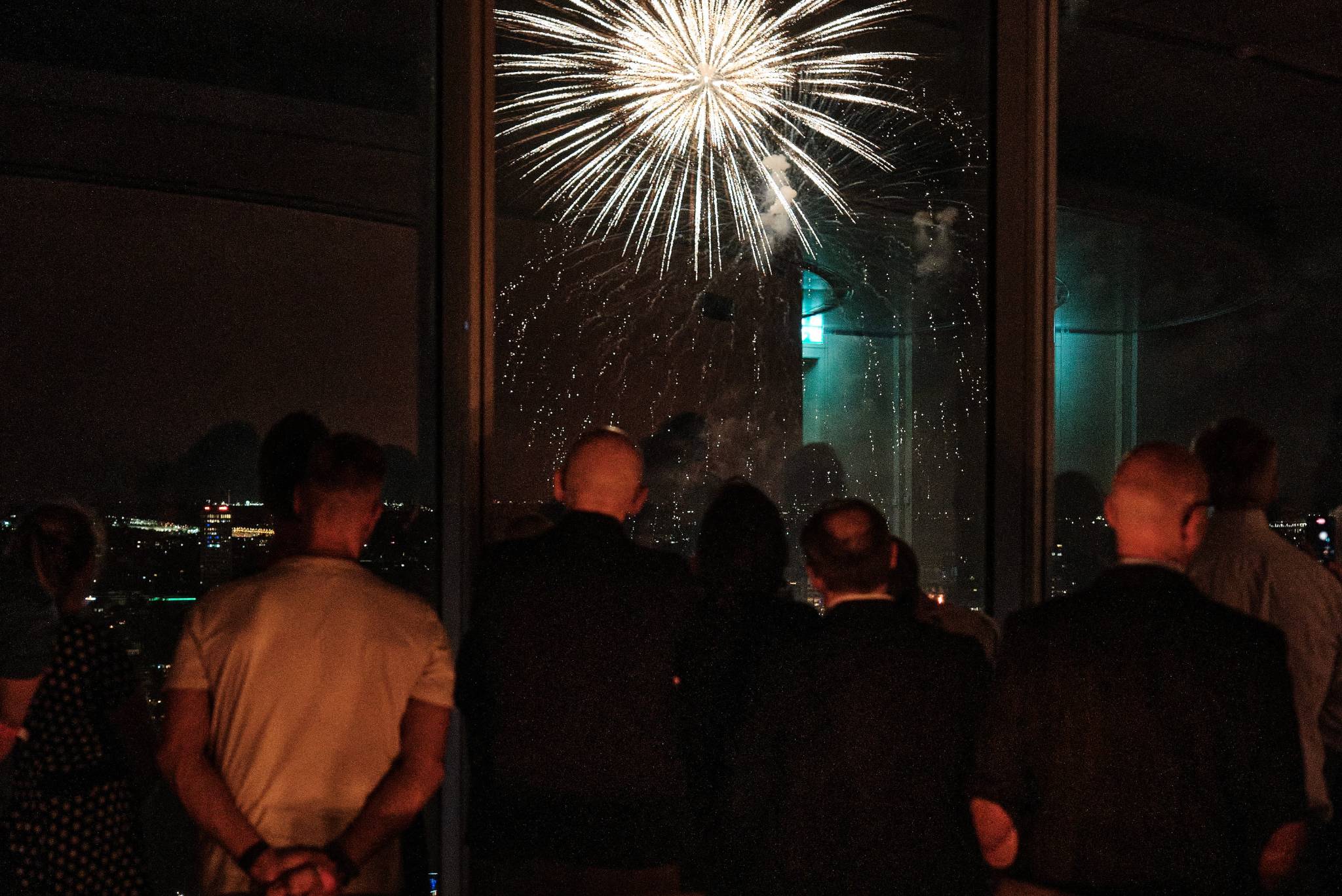 Im Inneren des KölnSky stehen die Gäste an den Fenstern und beobachten das Feuerwerk Spektakel, welches ebenfalls im Hintergrund zu sehen ist.